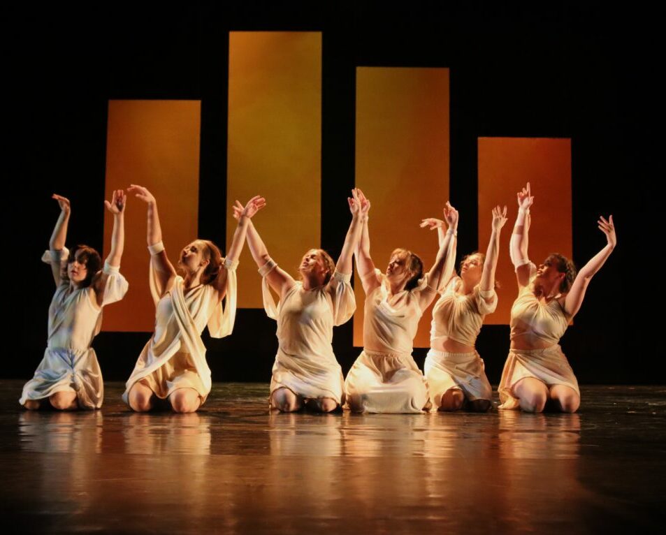 Photograph of the dance "Icarus and Apollo" from AFTERIMAGES 2023. Six dancers are in frame. They are kneeling with their arms raised. Behind them are four flats of different sizes hanging at different heights, which are painted in yellow and orange to look like a sunset. The background is completely black.